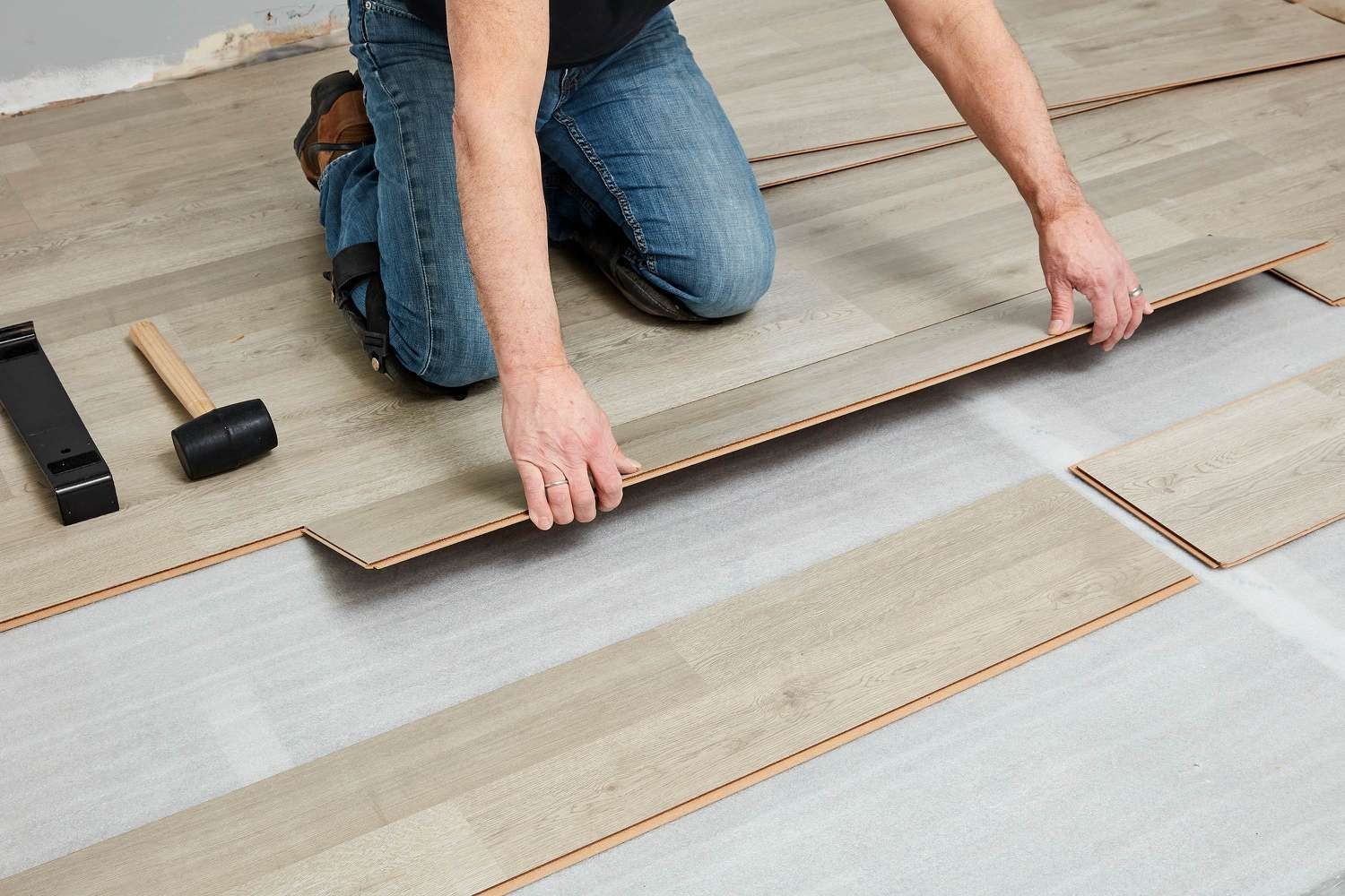 Person installing wooden laminate flooring in a room, using tools including a mallet and tapping block.